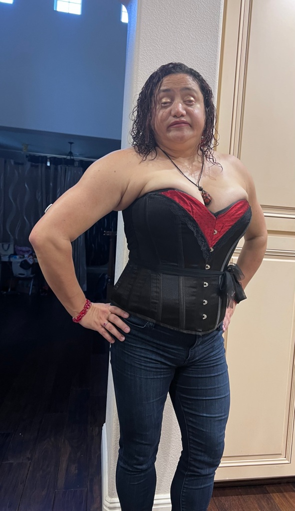 TheBlindWriter poses with her hand on her hip. She is wearing jeans and a black and red corset that emphasizes her large breasts. She is wearing her dark hair down and has on makeup.`